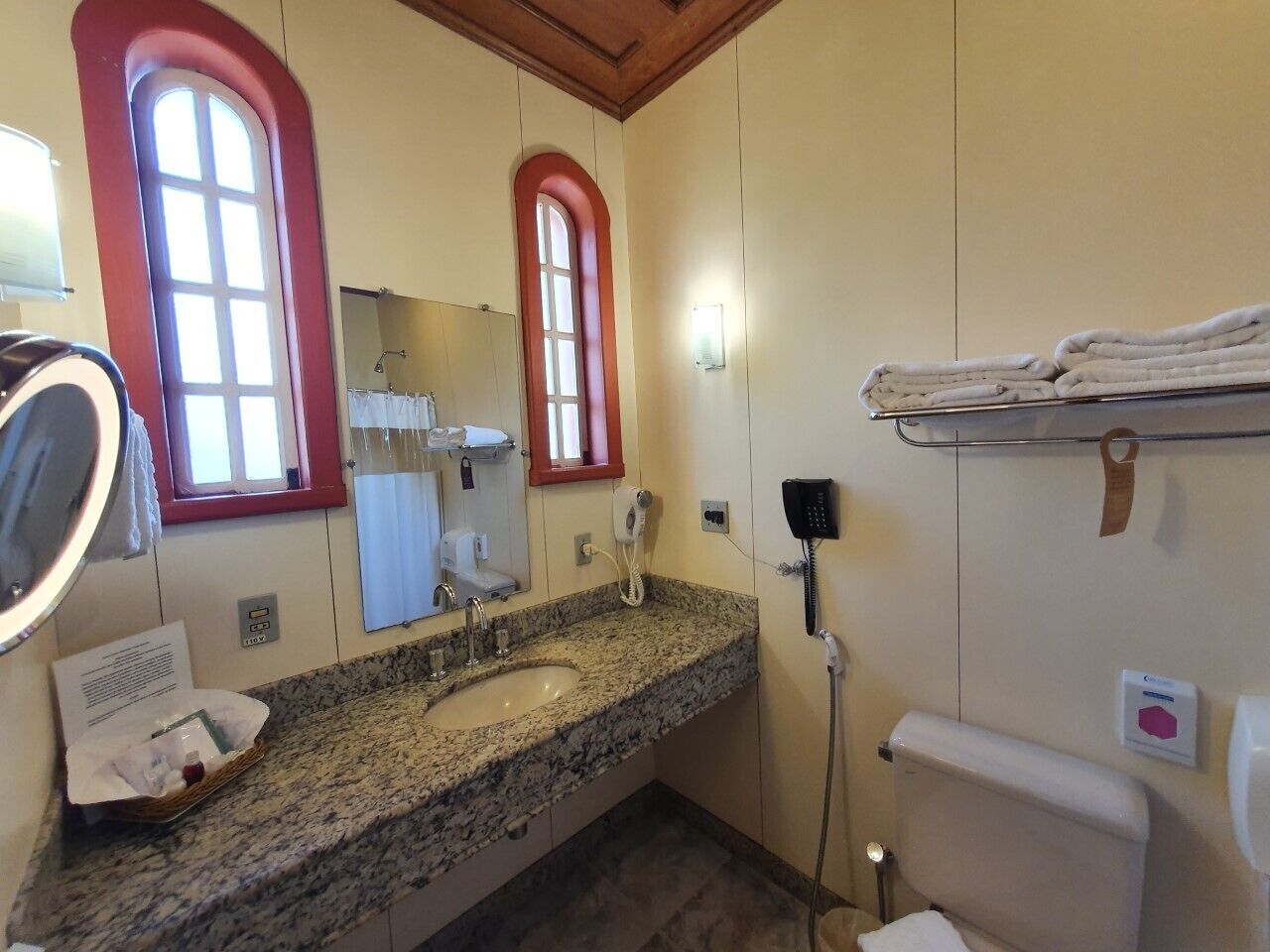 A bathroom with a granite sink, a mirror, a red-framed window, a towel rack, and a toilet.