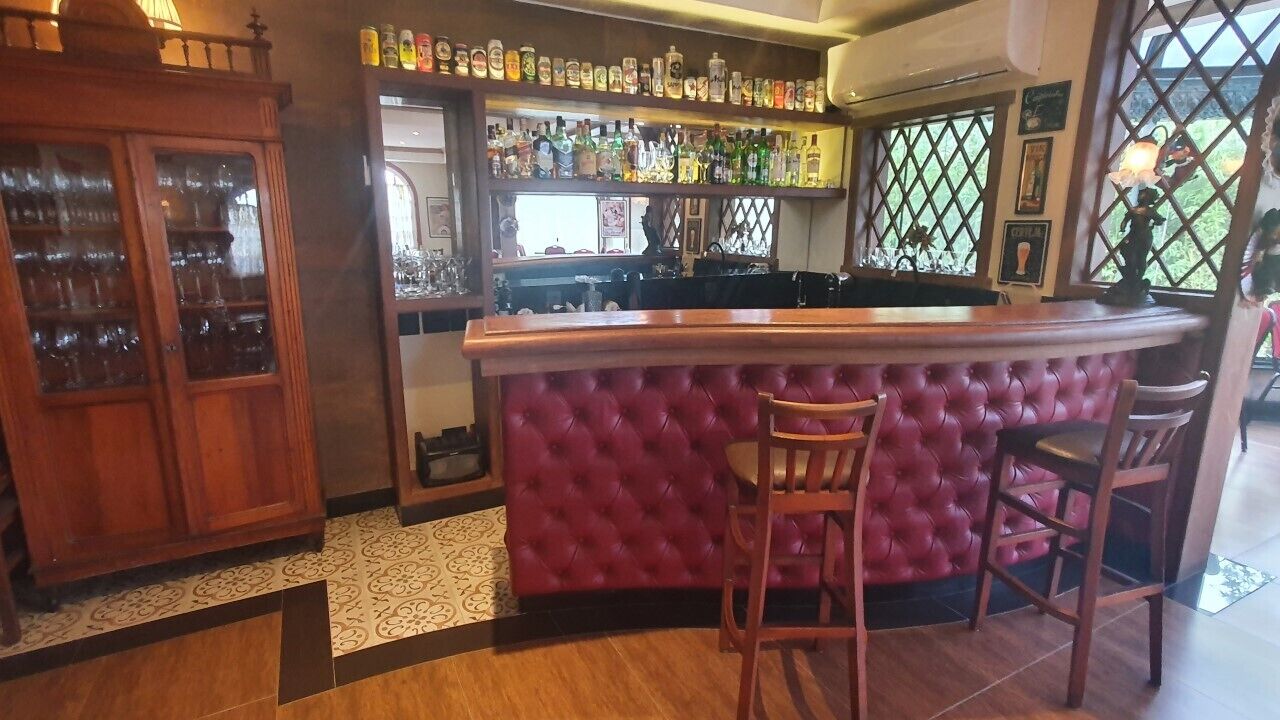 A bar area with a tufted bar counter and stools, a wooden cabinet with glass doors, and shelves with bottles and glasses.
