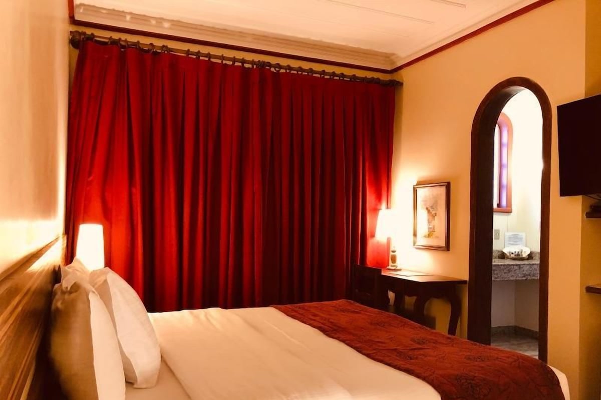 A hotel room with a bed, a desk, a chair, a lamp, and a mirror.