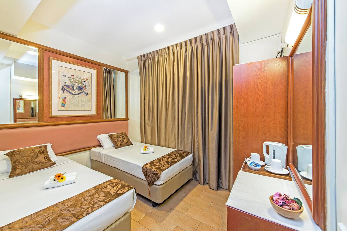 Quarto de hotel com duas camas, um guarda-roupa de madeira, um espelho, um quadro na parede e uma mesa pequena com chaleira e chávenas.