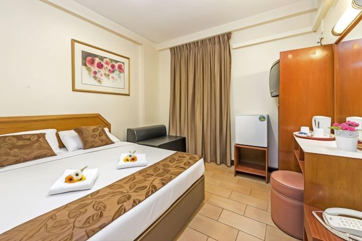 A hotel room with a bed, bedside table, a desk with a chair, a mirror, and a wardrobe.