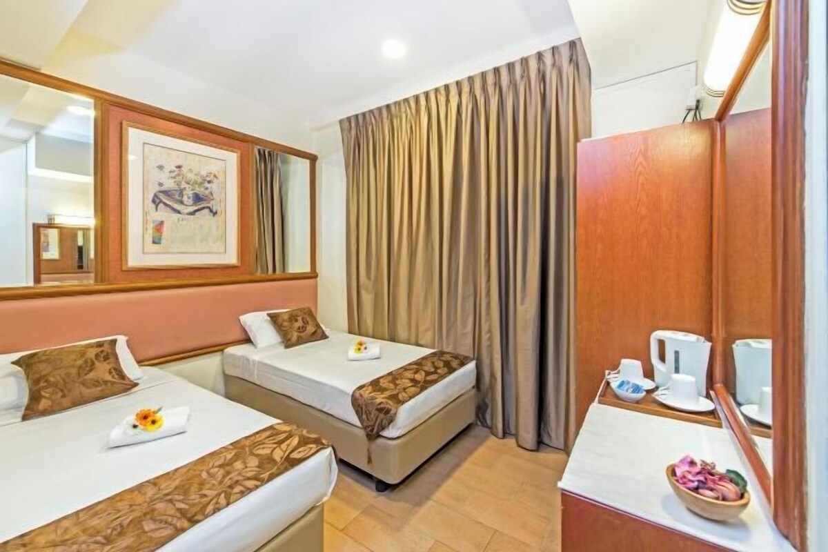 A hotel room with two beds, a wooden wardrobe, a mirror, a framed picture on the wall, and a small table with a bowl of fruit.