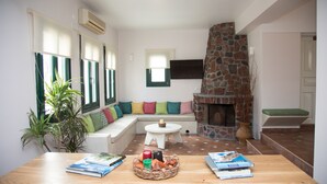 A living room with a stone fireplace, a white sofa with colorful cushions, a small white table, and a wooden table with books and a basket of fruit. - Costa Marina Villa (Santorini)