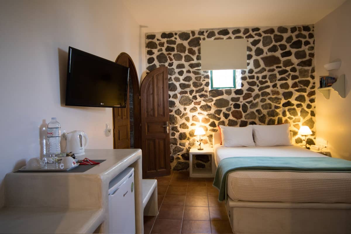 A small, cozy room with a stone wall, a bed with a green blanket, a bedside table with a lamp, a TV on the wall, and a kitchenette area with a coffee maker and water bottles.
