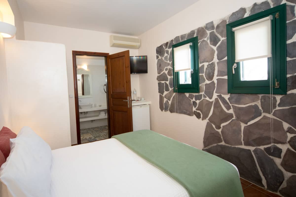 A bedroom with a bed, a green blanket, a stone wall, a television, and a bathroom visible through an open door.