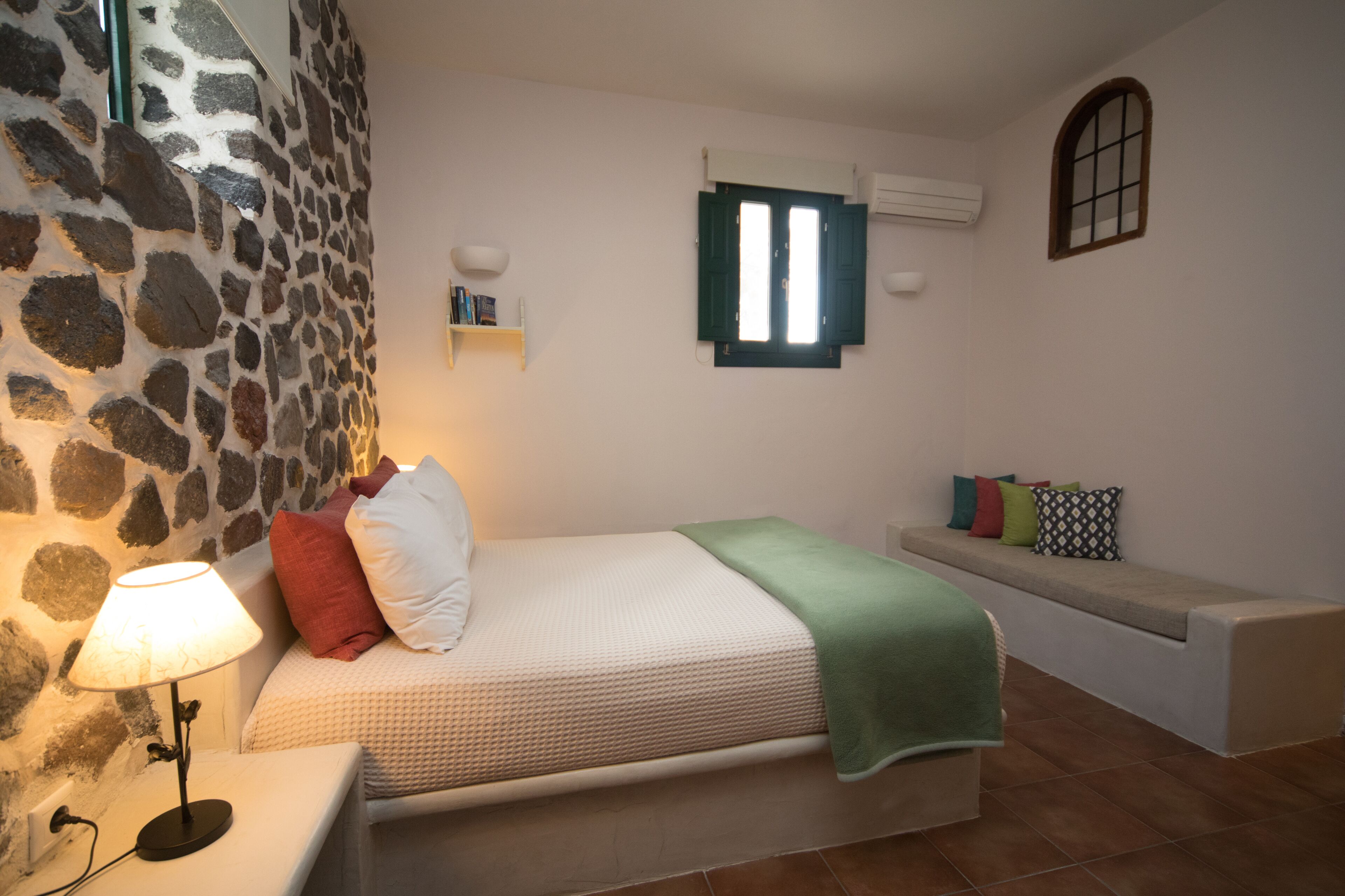 A bedroom with a stone wall, a bed with pillows, a bedside table with a lamp, a bench with cushions, and a window with shutters.