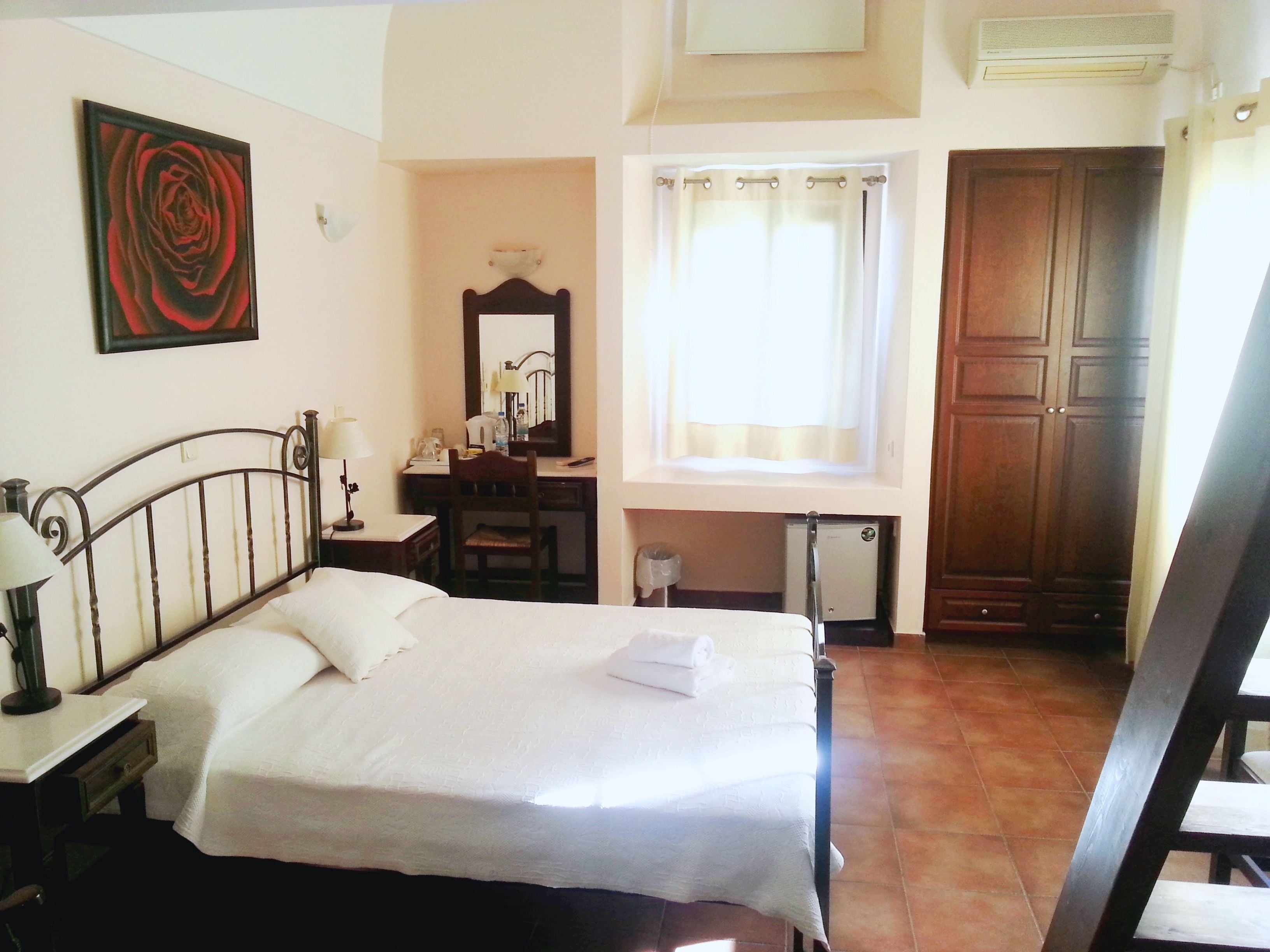 Une chambre d’hôtel comprenant un lit, un bureau, une chaise, un miroir, une fenêtre avec des rideaux et une armoire en bois.