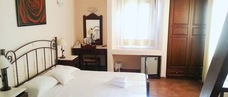 Une chambre d’hôtel comprenant un lit, un bureau, une chaise, un miroir, une fenêtre avec des rideaux et une armoire en bois.