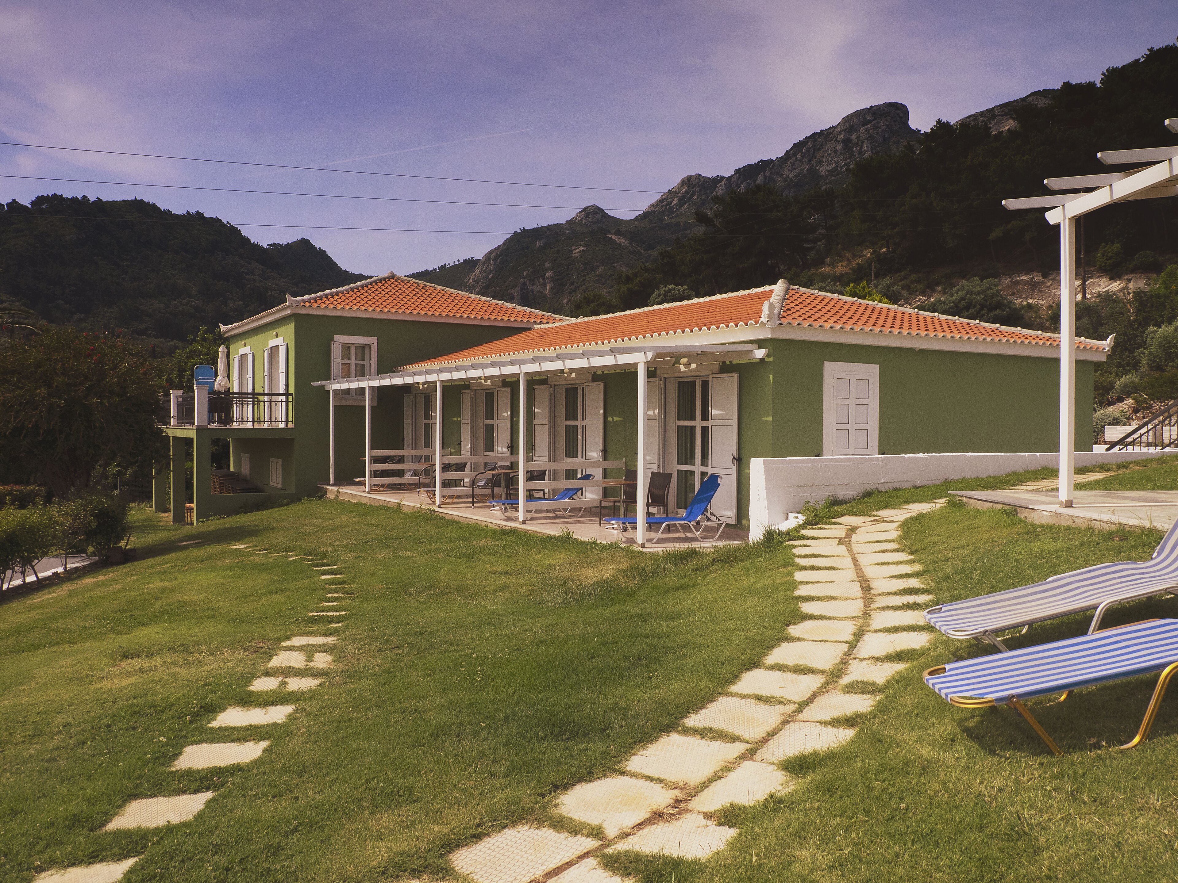 A green villa with a tiled roof, a covered patio with chairs and umbrellas, and a stone pathway leading to the entrance.