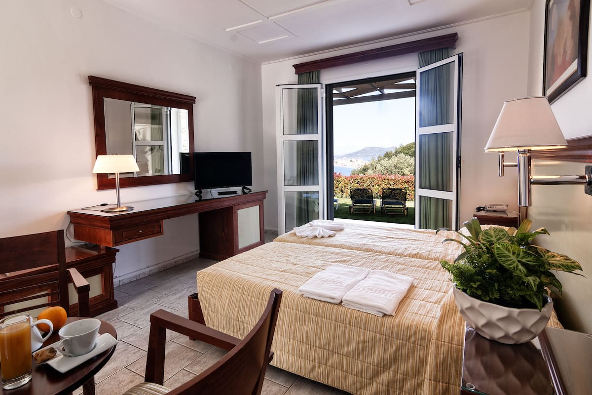 A hotel room with a bed, a desk with a TV, a chair, a table with a cup and a glass, and a window with a view of the outdoors.