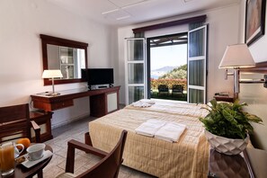 A hotel room with a bed, a desk with a TV, a chair, a table with a cup and a glass, and a window with a view of the outdoors.