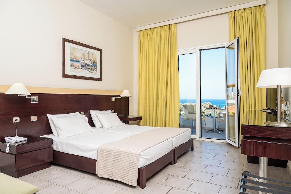 A hotel room with a large bed, a bedside table with a lamp, a small table with a lamp, a painting on the wall, and a balcony with yellow curtains.