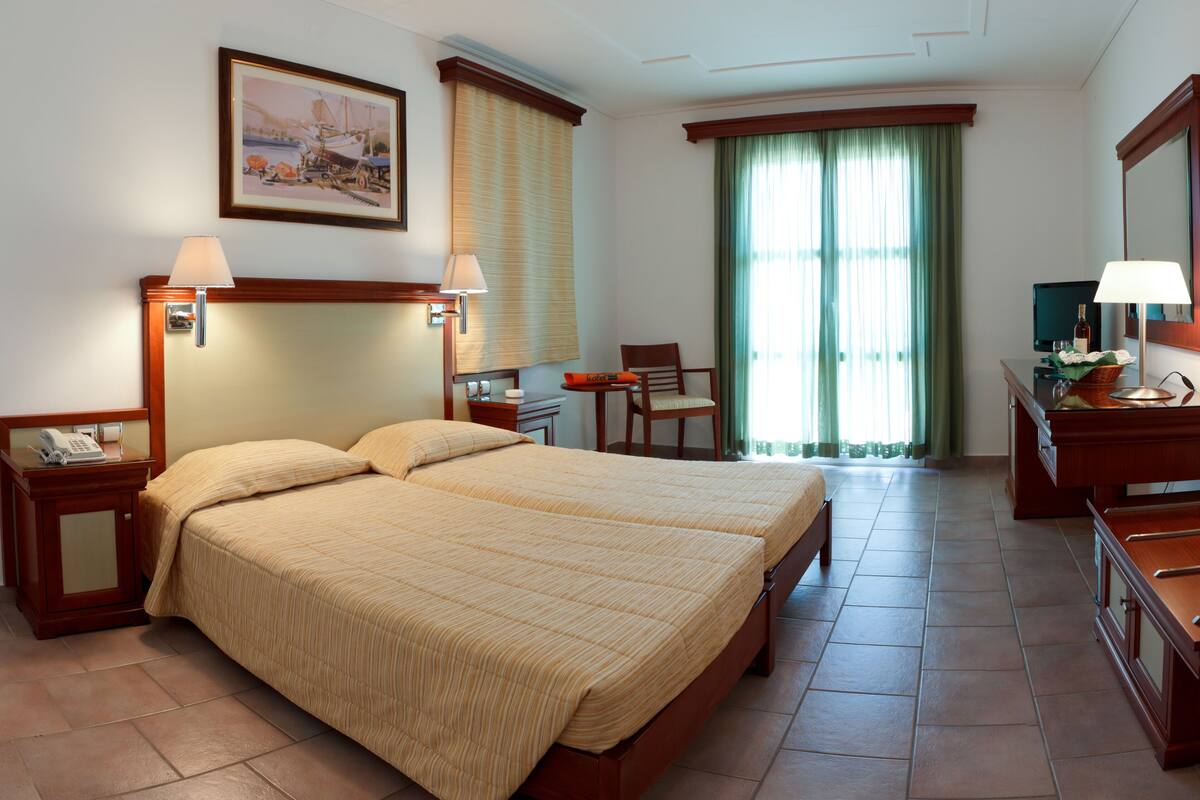 A hotel room with a double bed, a desk, a chair, a telephone, a lamp, a mirror, and a painting on the wall.