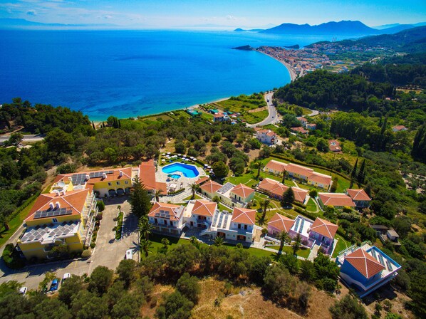 An aerial view of a coastal residential area with a swimming pool, multiple houses, and a road leading to the sea. - Arion Hotel (Samos)