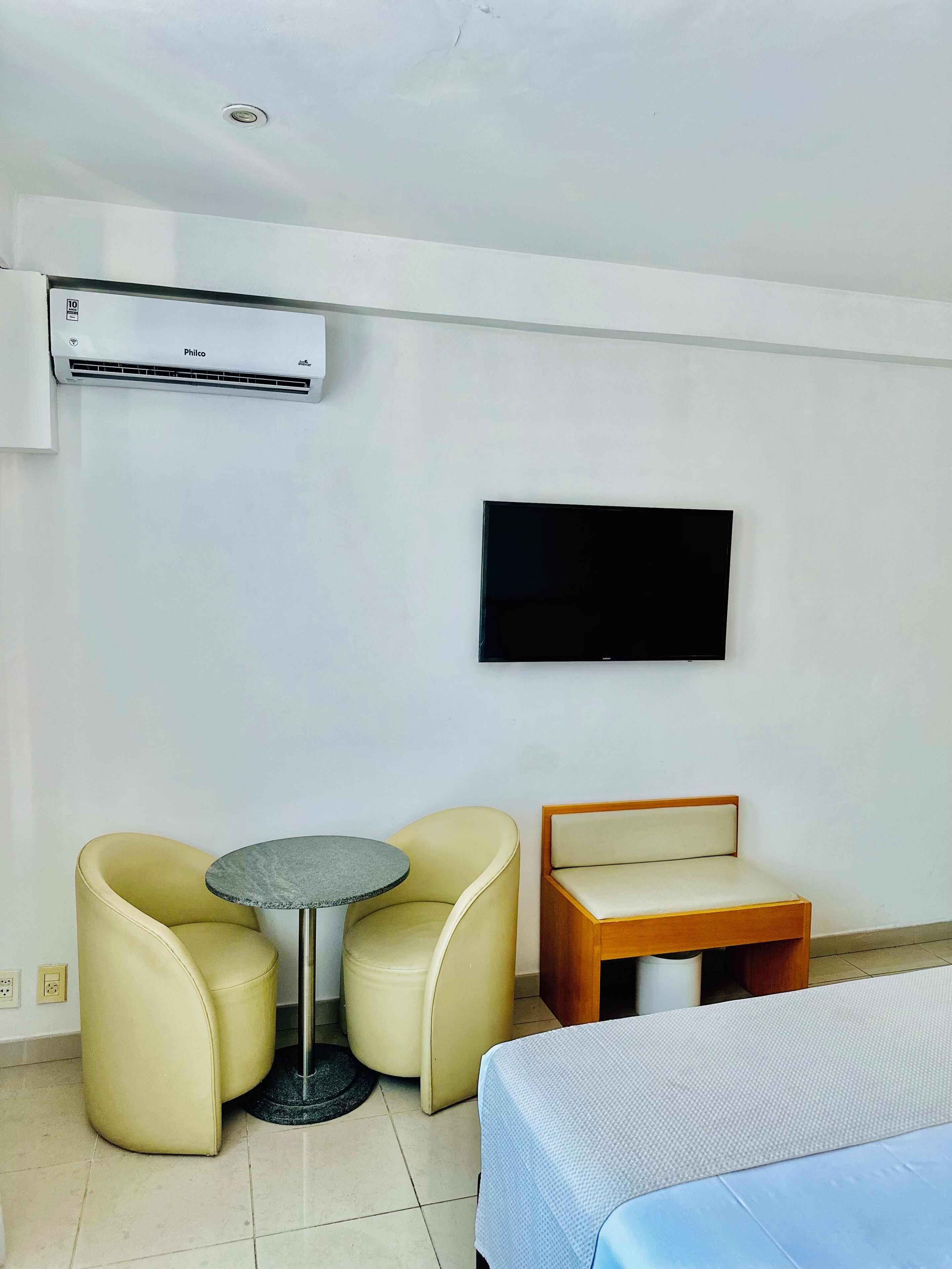 A hotel room with a bed, a TV mounted on the wall, a small round table, two armchairs, and an air conditioning unit.