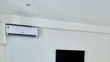 A hotel room with a bed, a TV mounted on the wall, a small round table, two armchairs, and an air conditioning unit.