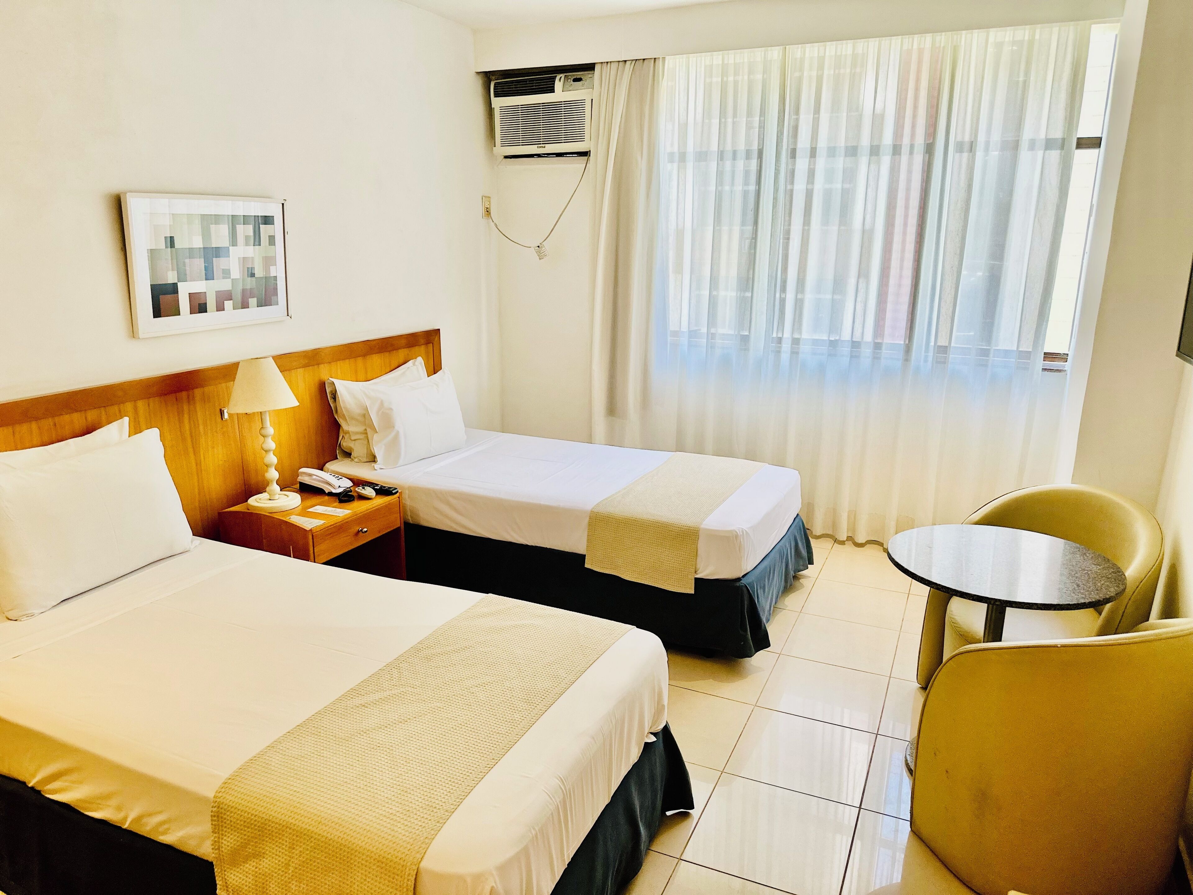 Quarto de hotel com duas camas, uma mesa de cabeceira com abajur, uma cadeira e uma pequena mesa.