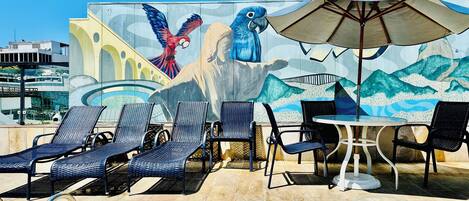 Un área de estar al aire libre con sillones reclinables y una mesa, un mural con un loro y montañas, y un cielo azul despejado.