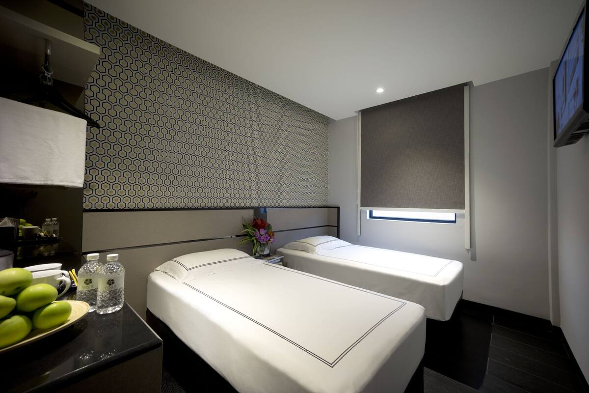 Une chambre d’hôtel avec deux lits, une petite table sur laquelle est posé un vase de fleurs, un téléviseur fixé au mur et un mur décoré d’un motif.