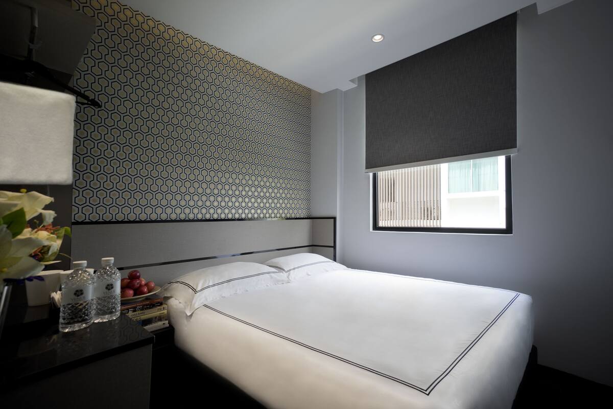 Une chambre à coucher moderne avec un mur orné d’un motif géométrique, une grande fenêtre avec des stores, un lit soigneusement fait et une table de chevet agrémentée d’un vase et de livres.