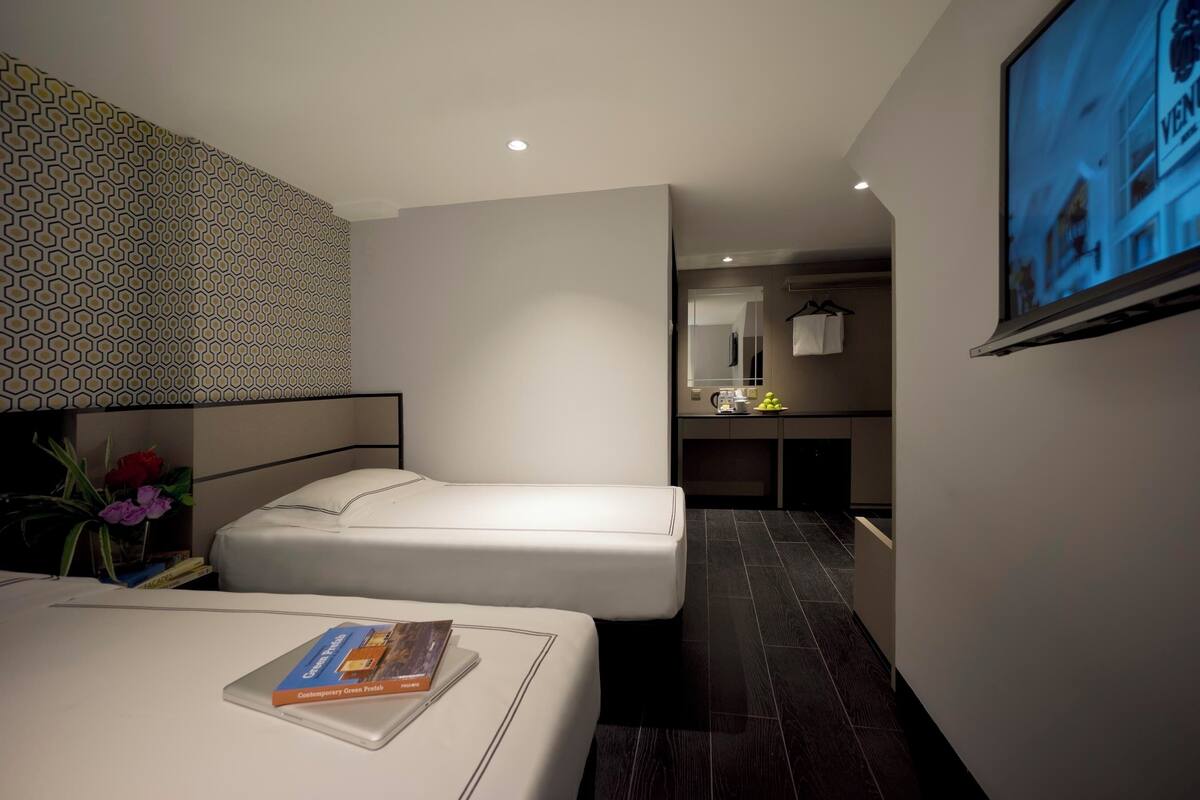 Une chambre d’hôtel moderne avec deux lits, une petite kitchenette, un miroir et un téléviseur fixé au mur.