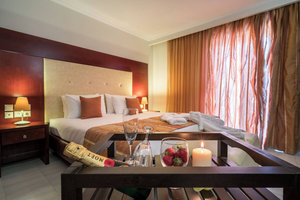 A hotel room with a large bed, a bedside table with a lamp, a glass of water, a bottle of champagne, and a candle.