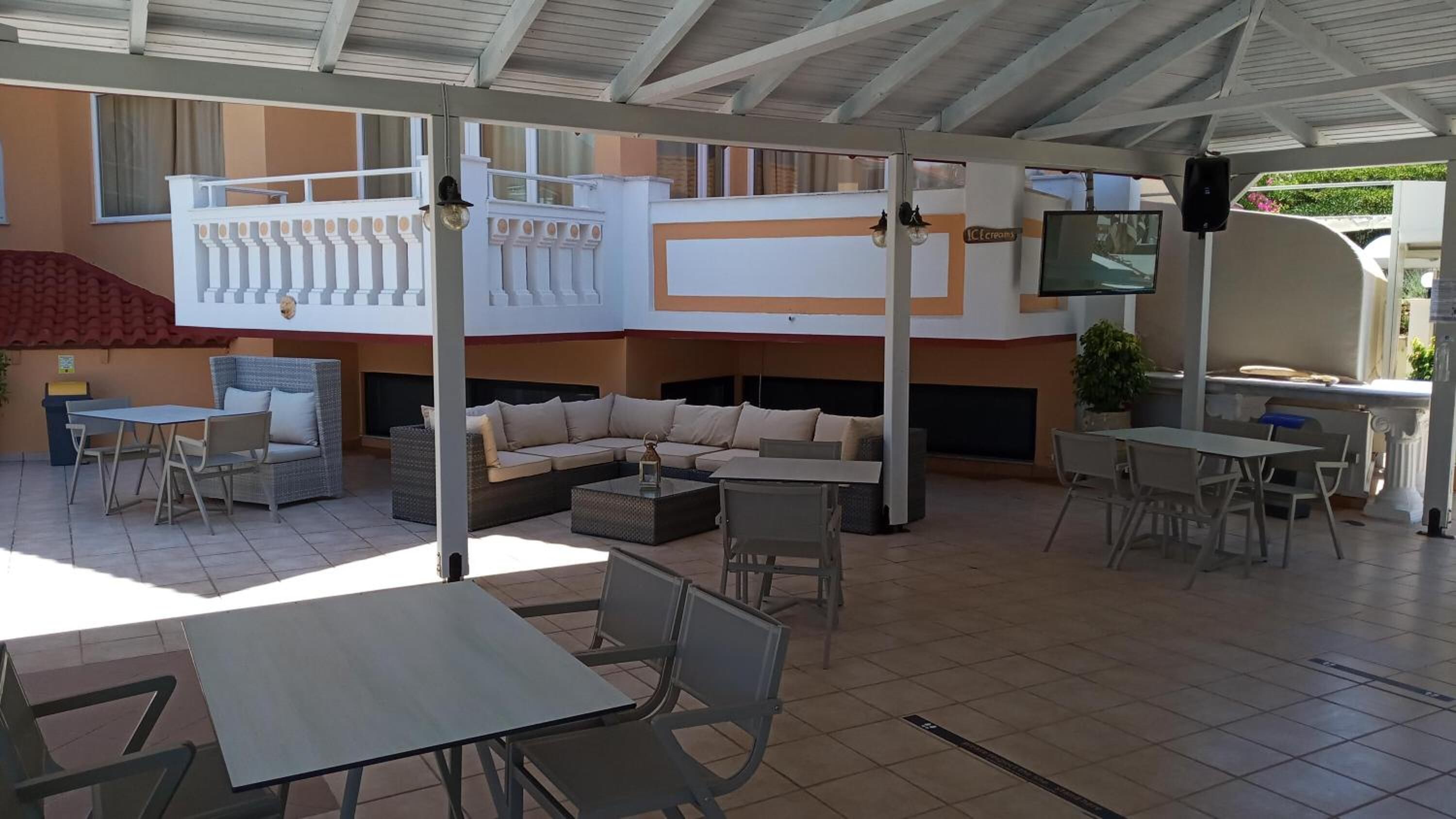 A covered outdoor seating area with tables and chairs, a sofa, and a TV screen.