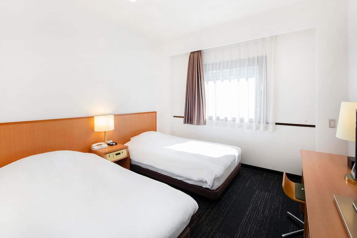 Une chambre d’hôtel avec deux lits, un bureau, une chaise, une lampe et une fenêtre avec des rideaux.