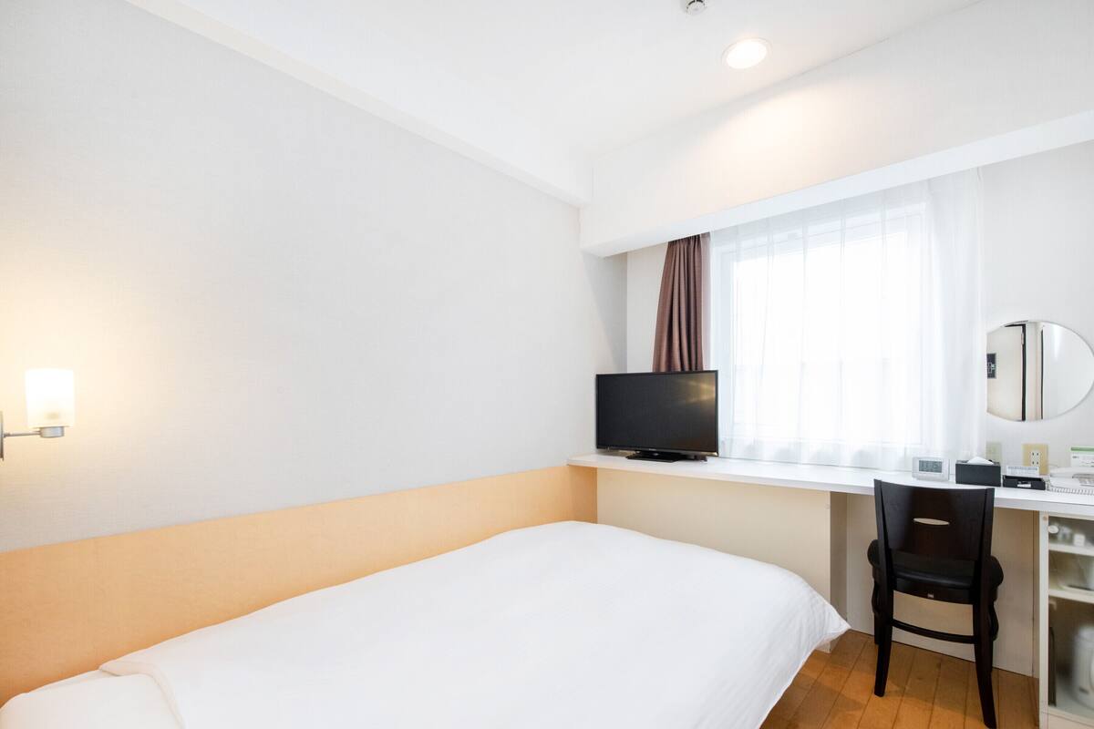 Une chambre d’hôtel avec un lit simple, un bureau avec une chaise, une télévision et une fenêtre avec des rideaux.