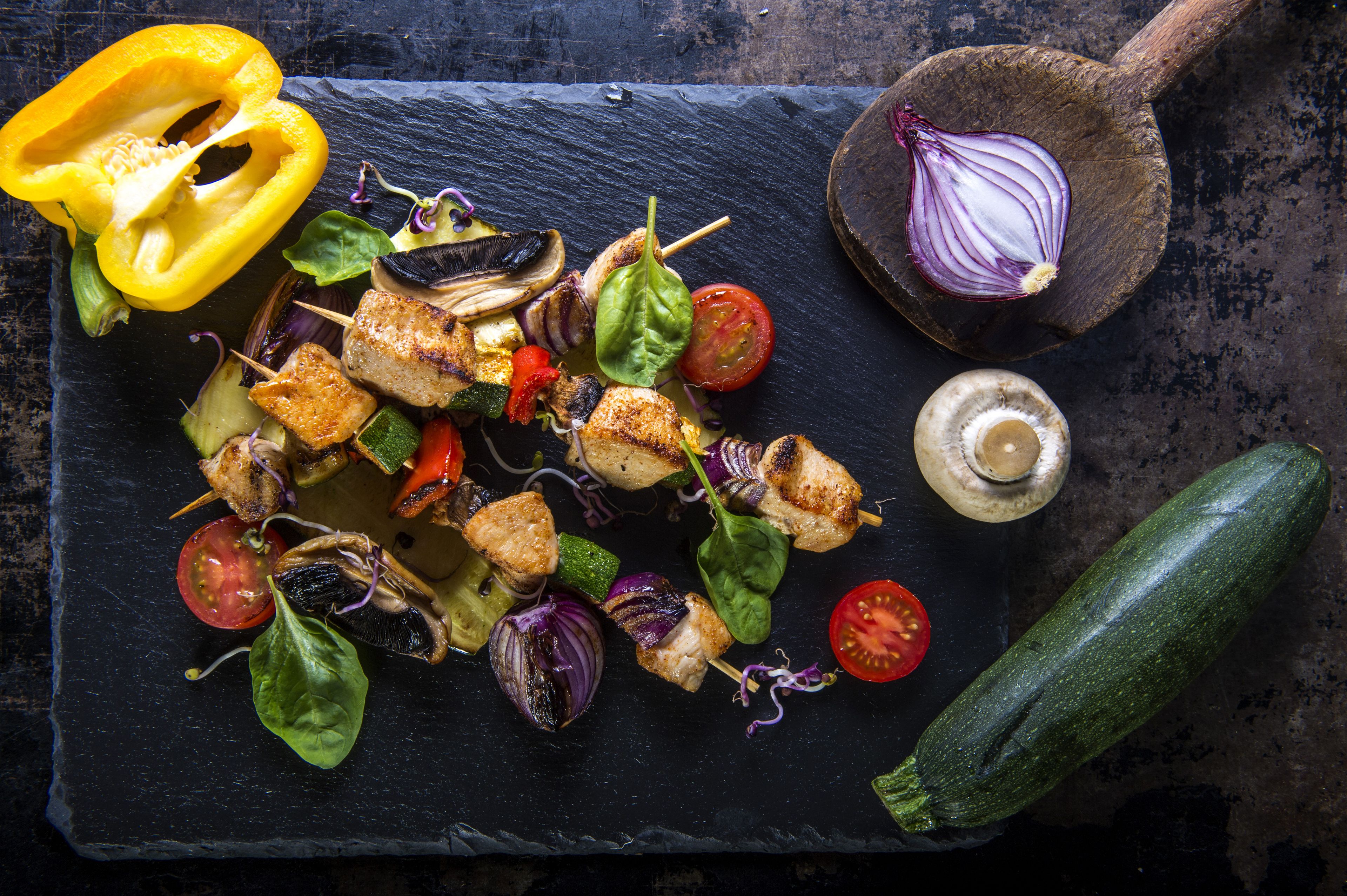 A variety of grilled vegetables and skewers on a slate board, including bell peppers, zucchini, and tomatoes.