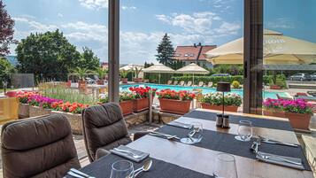 An outdoor dining area with tables set for two, overlooking a swimming pool and a garden with potted plants and flowers.