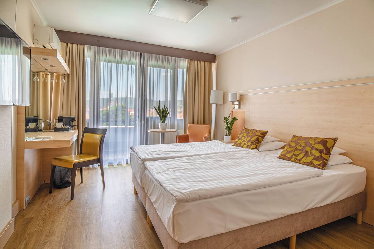 Una habitación de hotel con una cama grande, un escritorio con una silla, una mesita con una planta y una ventana con cortinas.