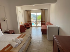 A hotel room with a bed, a desk, a chair, a sofa, and a view of a pool and greenery outside. - Stellina Hotel (Skiathos)