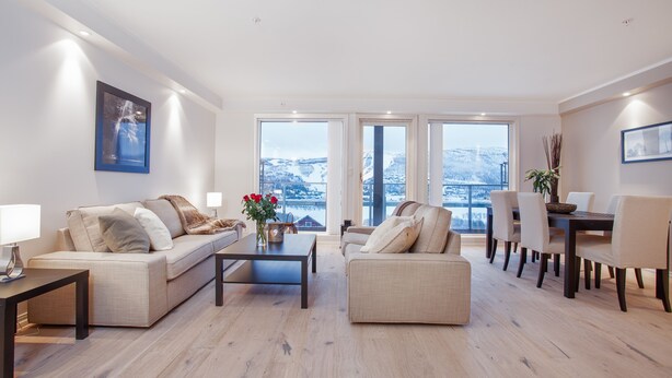 A modern living room with a sofa, coffee table, and dining area. There are large windows with a view of a snowy landscape. - Ustedalen Hotell Geilo (Hol)