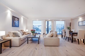 A modern living room with a sofa, coffee table, and dining area. There are large windows with a view of a snowy landscape. - Ustedalen Hotell Geilo (Hol)