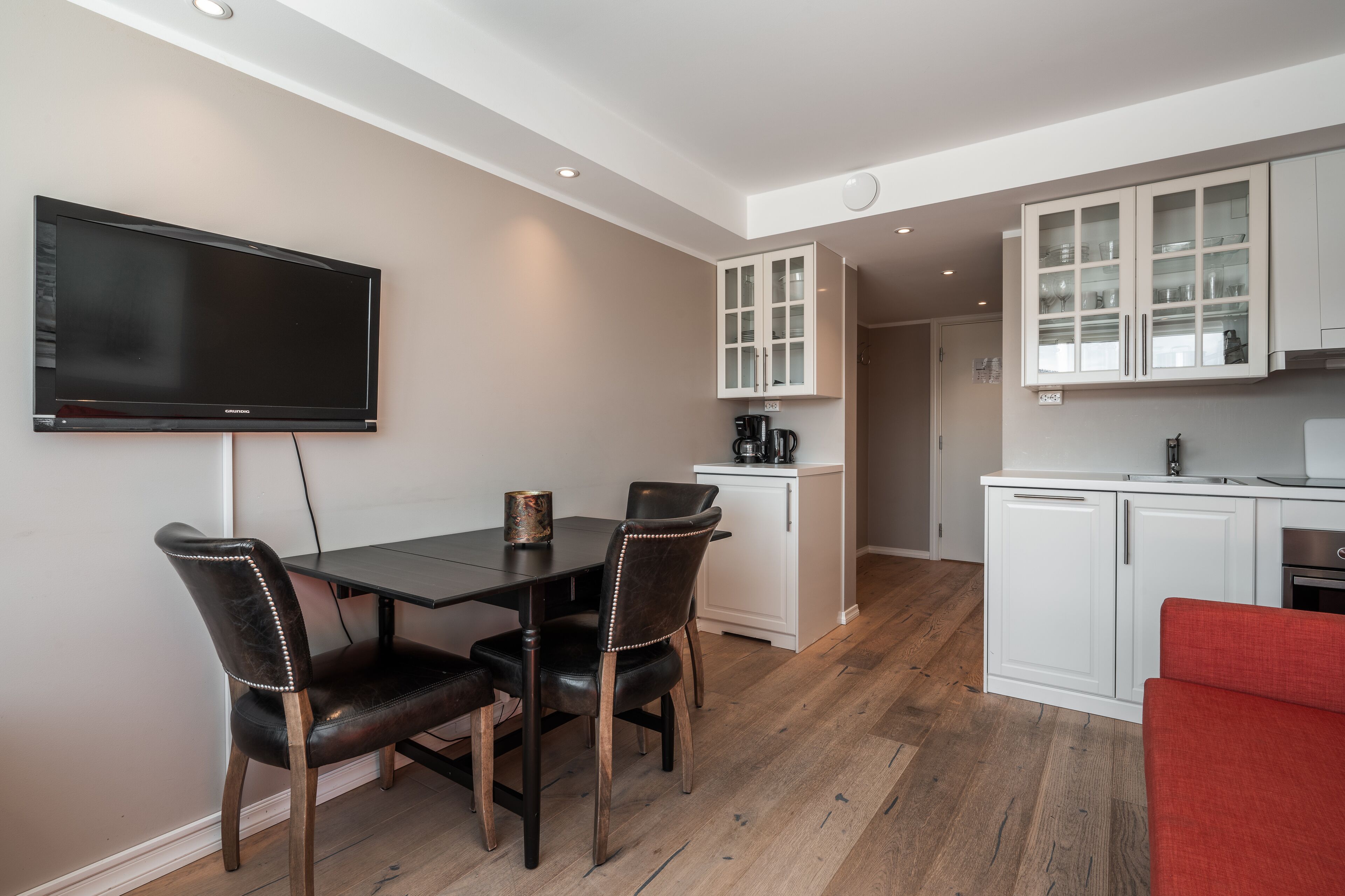 A modern kitchen with white cabinets, a dining area with a table and chairs, and a flat-screen TV mounted on the wall.