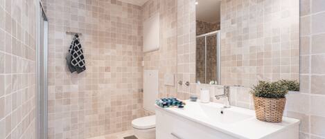 A modern bathroom with beige tiles, a glass shower enclosure, a white sink with a plant, and a toilet.