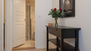 A hallway with a wooden floor, a black console table with a vase of red flowers, a framed picture on the wall, and a wicker basket with a candle.
