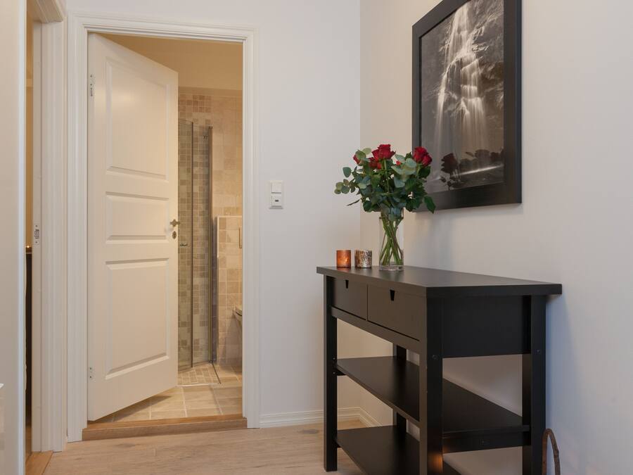 A hallway with a wooden floor, a black console table with a vase of red flowers, a framed picture on the wall, and a wicker basket with a candle.