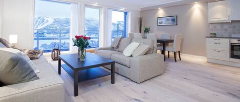 A modern living room with a sofa, a dining table, and a kitchen area. There are large windows providing a view of a snowy landscape.