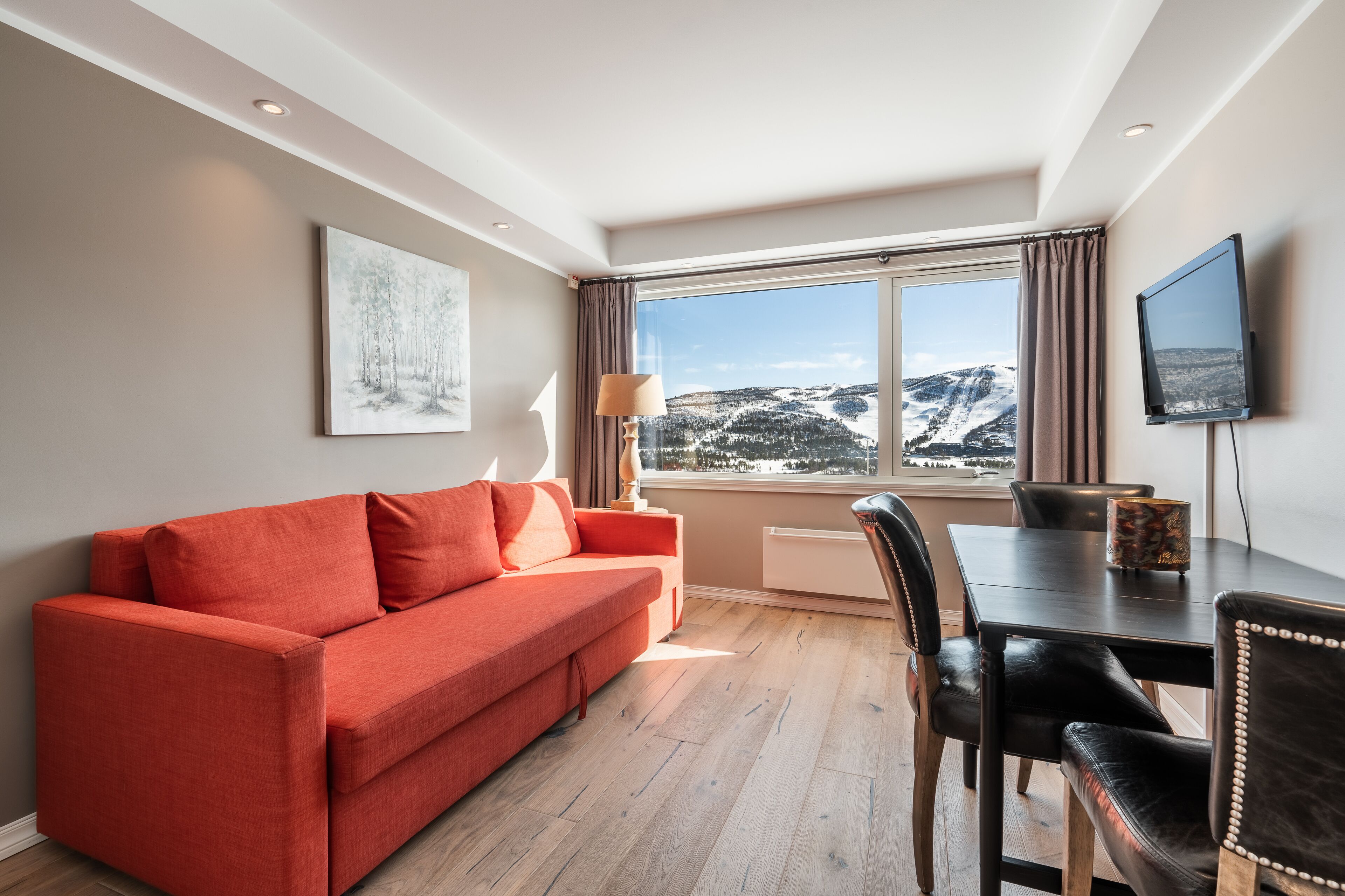A modern living room with a red sofa, a dining table with chairs, a TV, and a large window with a view of snow-covered hills.