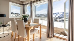 A dining area with a wooden table and chairs, large windows with curtains, and a view of a snowy landscape. - Ustedalen Hotell Geilo (Hol)