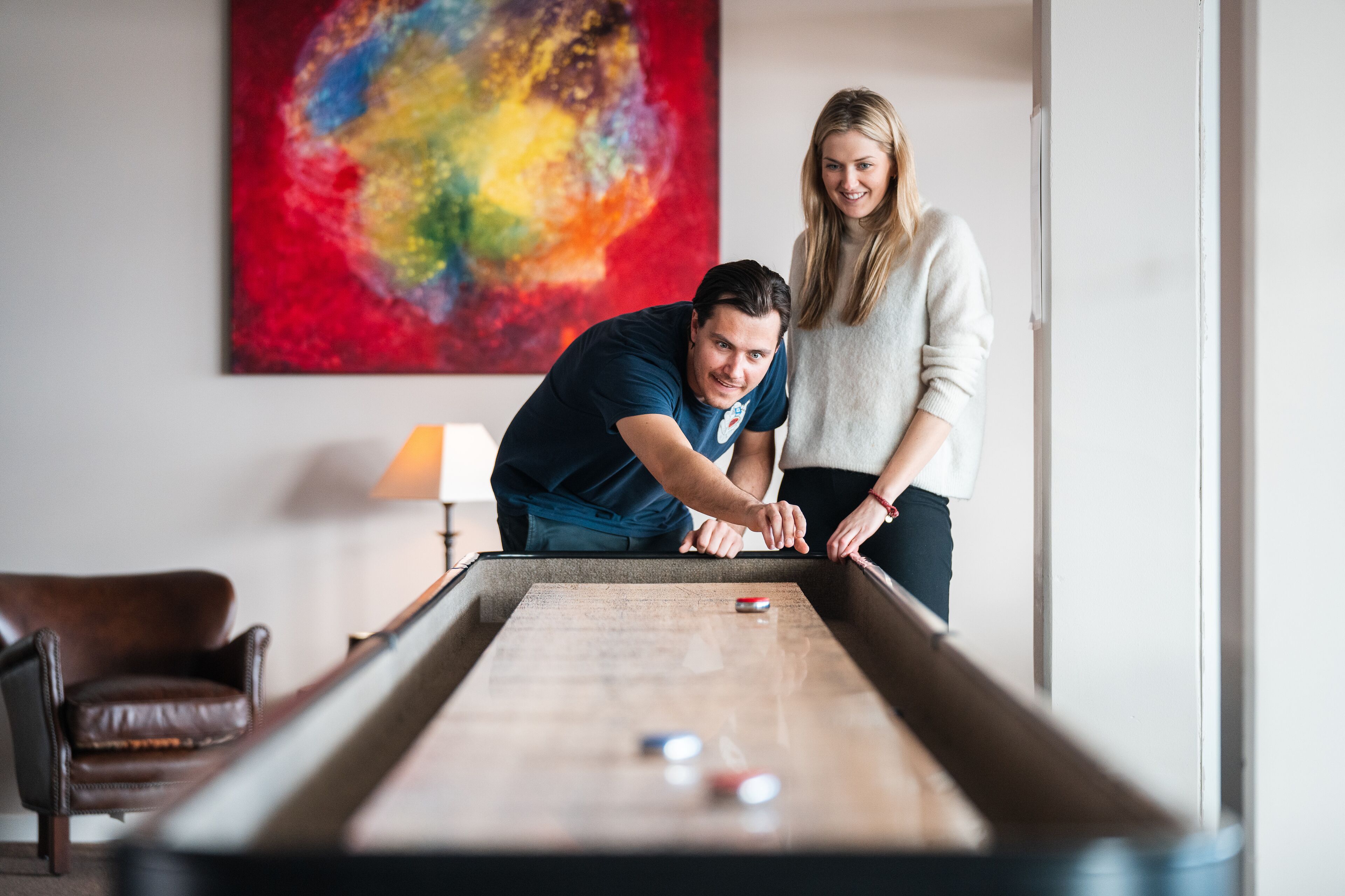 A pool table with two people playing, a brown leather chair, and a colorful abstract painting on the wall.