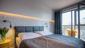 A modern bedroom with a brick wall, a large bed, a bedside table with a vase of flowers, and a window with a view of trees. - Hotel Czarna Róża (Torun)