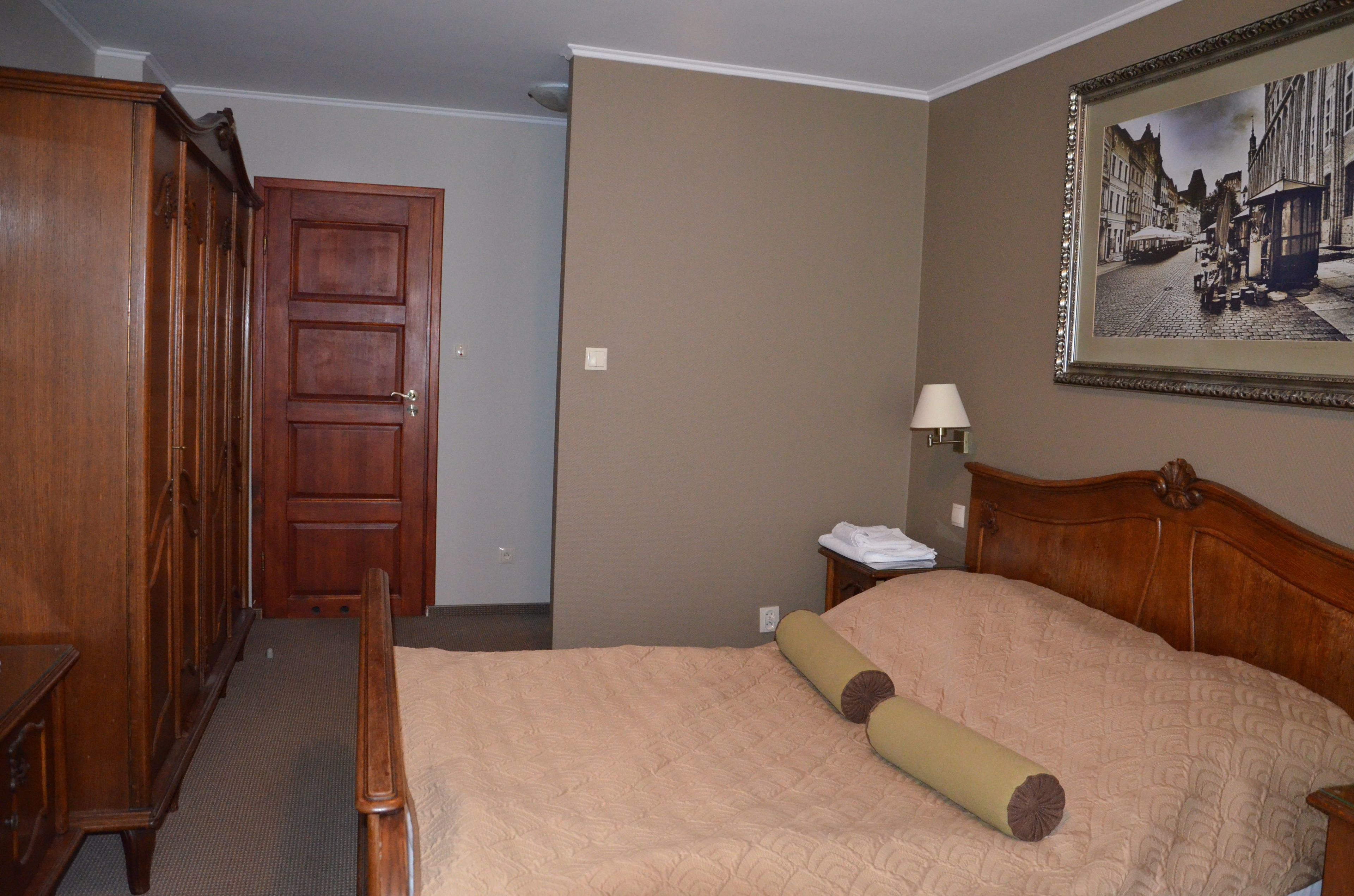 Een hotelkamer met een houten bed, een nachtkastje met een lamp, een lijst met een foto aan de muur en een houten garderobe.