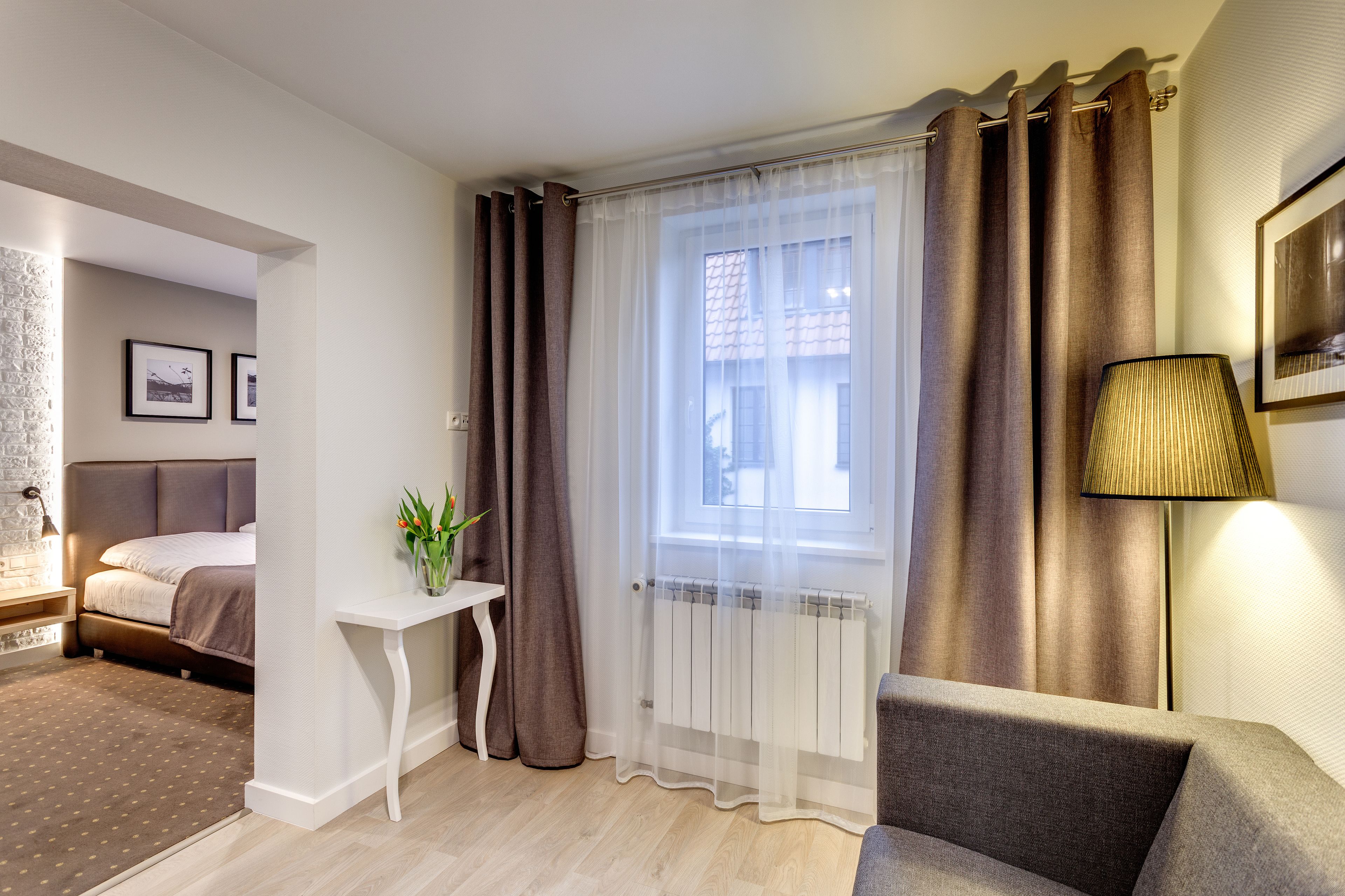 Habitación de hotel con cama, una mesita con una planta, ventana con cortinas, radiador y lámpara.