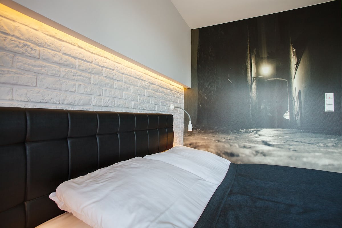 A modern bedroom with a brick wall, a black upholstered headboard, and a large painting of a dark corridor.