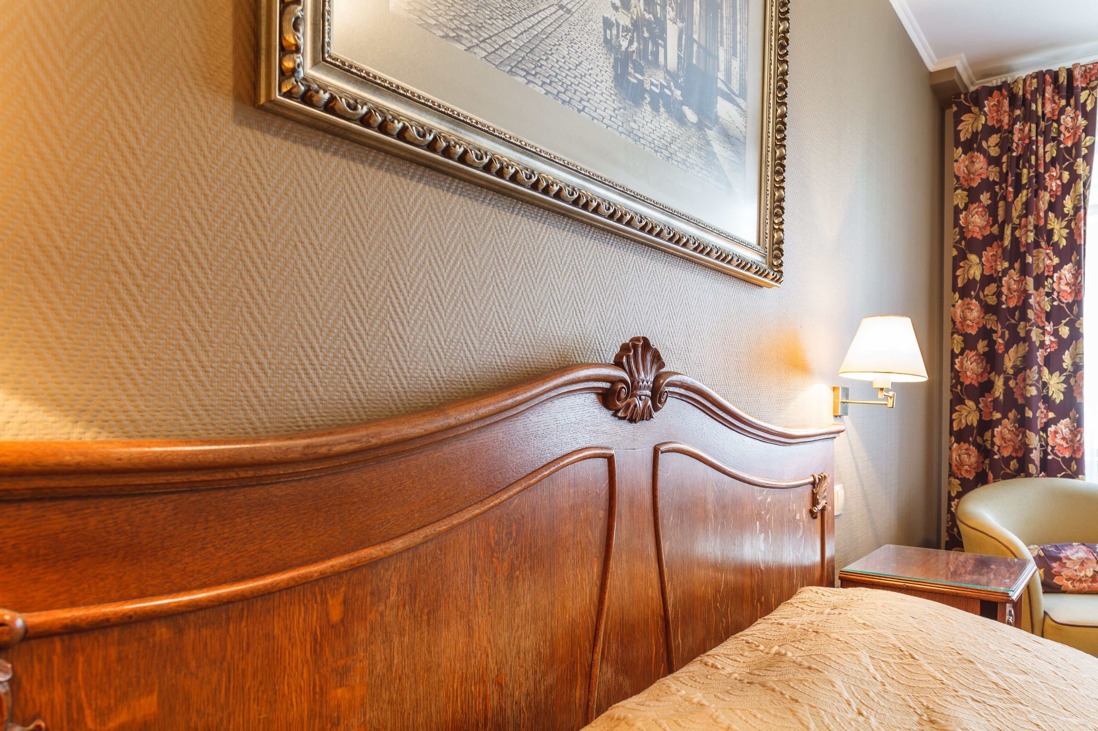 Una habitación de hotel con cabecera de madera, un cuadro en la pared, una mesita de noche y una cortina con estampado floral.