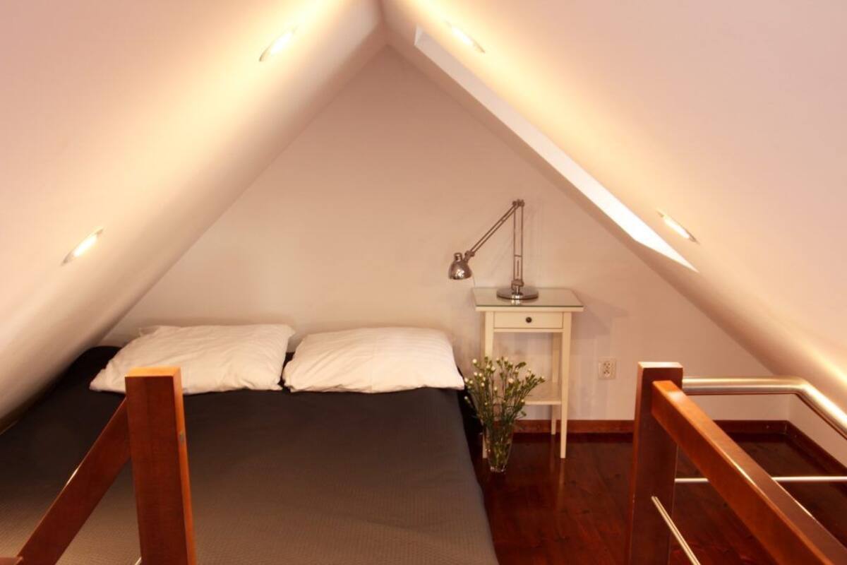 A small, triangular attic bedroom with a bed, pillows, a nightstand, a lamp, and a small plant.