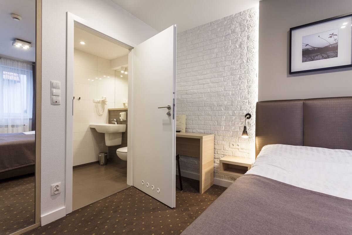 A hotel room with a bed, a bedside table, a nightstand, a desk, a chair, a mirror, a sink, and a toilet.